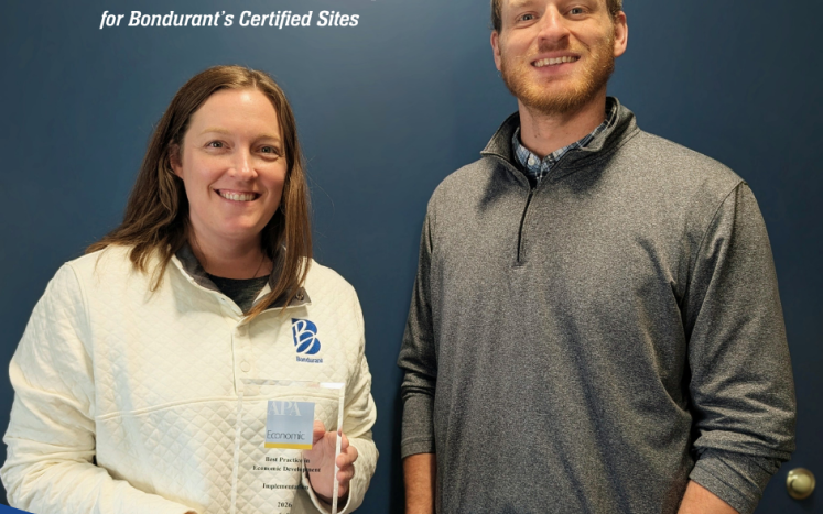 Two City of Bondurant staff members stand smiling in front of a blue wall, one holding an award plaque.